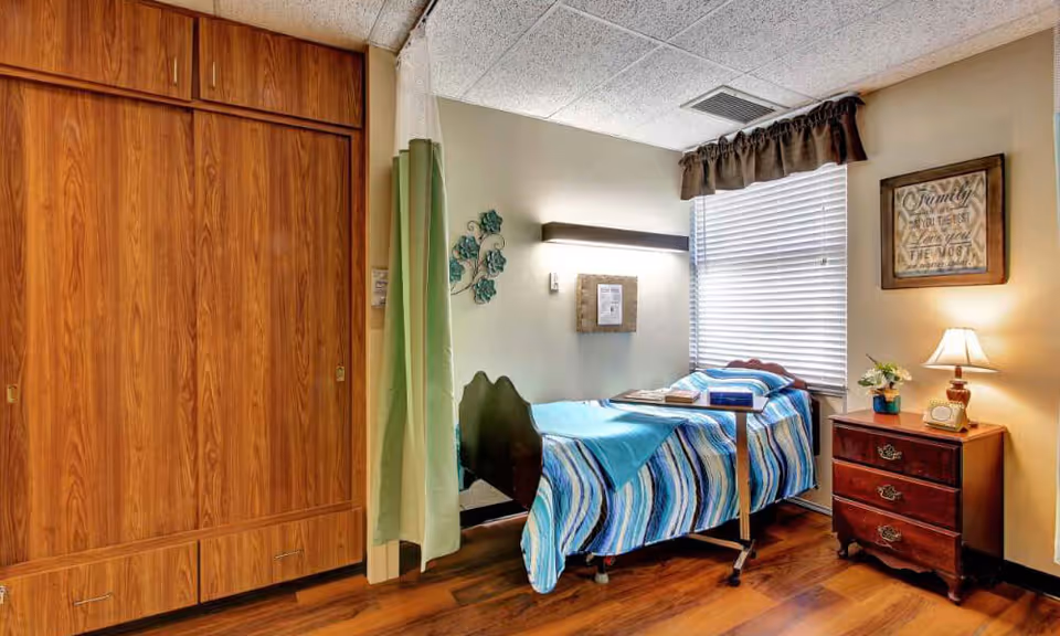 A small, cozy bedroom in a senior living facility with a single bed covered in a blue striped blanket. Next to the bed is a wooden nightstand with a lamp, a small plant, and a clock. The room has a large wooden closet with sliding doors, a window with blinds and a valance, and a wall decoration with a framed inspirational quote. The floor is wooden, and there is a privacy curtain partially drawn beside the bed.