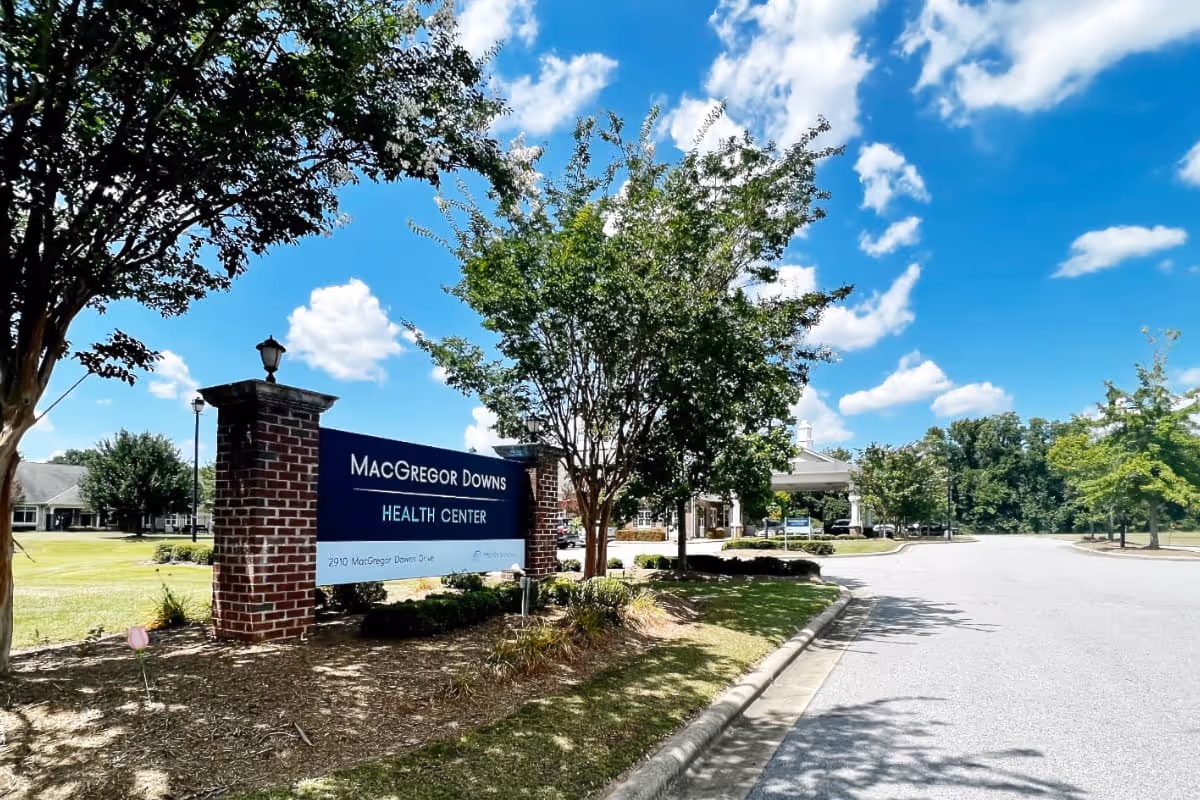 Exterior view of MacGregor Downs Health Center with a brick sign displaying the facility name and address. The scene includes a paved driveway, green trees, and a bright blue sky with scattered clouds.