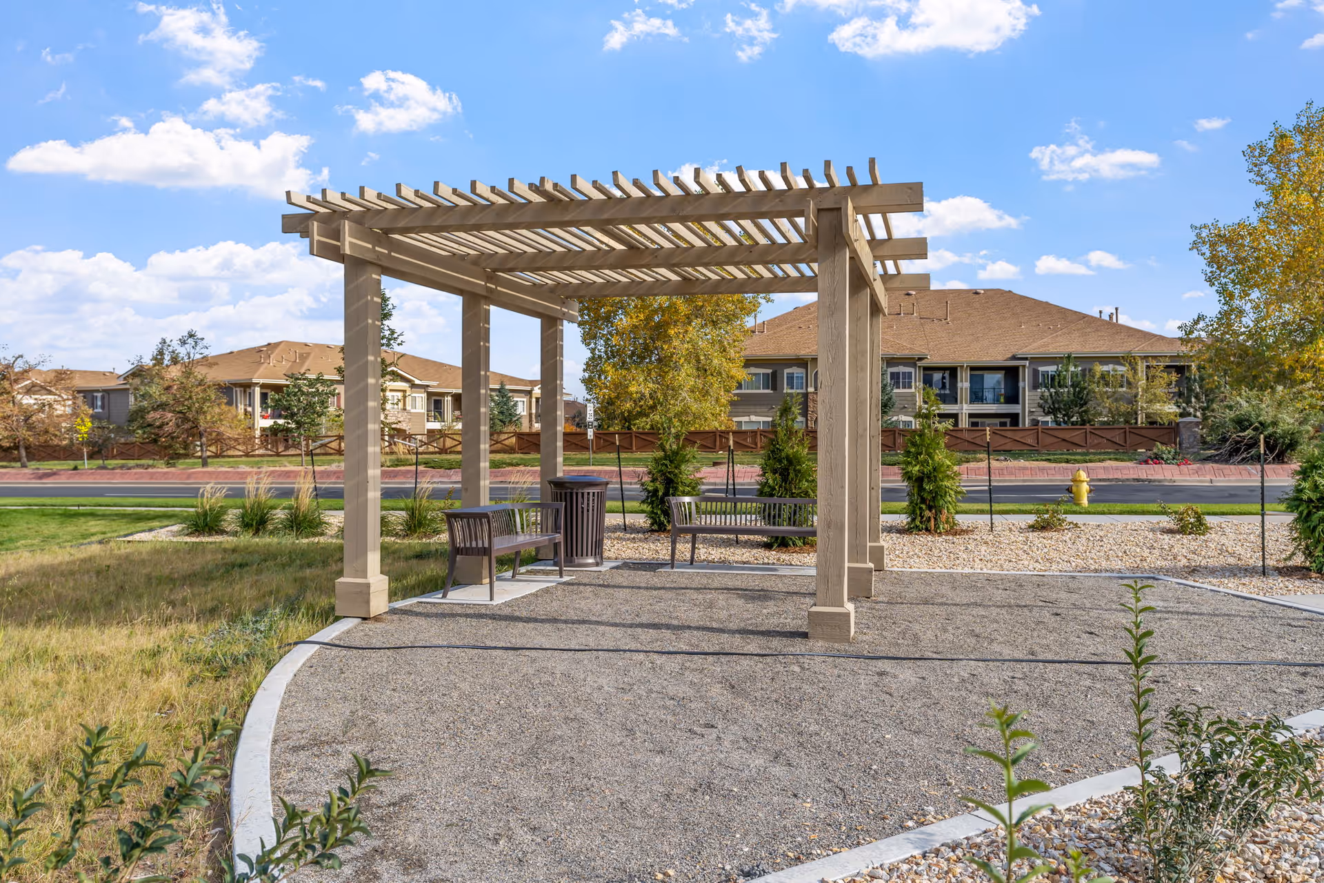 Outdoor seating area with a wooden pergola providing partial shade over two benches and a trash can, surrounded by gravel and landscaping with grass, shrubs, and trees. Residential buildings and a street with a yellow fire hydrant are visible in the background under a partly cloudy sky.