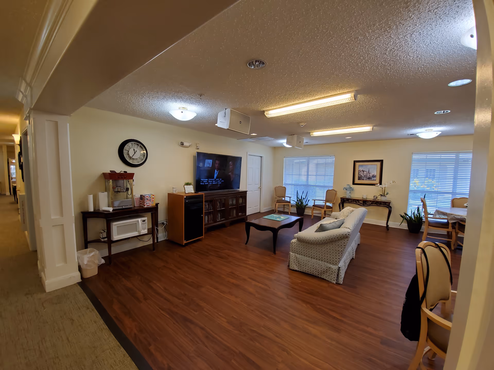 A spacious lobby area with a comfortable couch, a television, and dining tables. The room features wooden flooring, potted plants, and large windows with blinds.