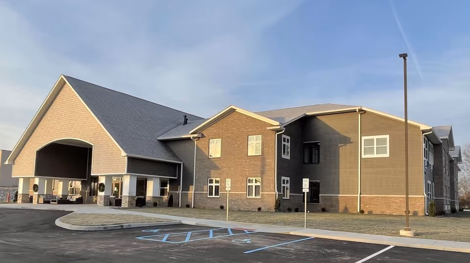Exterior view of a two-story senior living facility building with a covered entrance, multiple windows, and a parking lot with handicap spaces in front. The sky is clear with some light clouds.