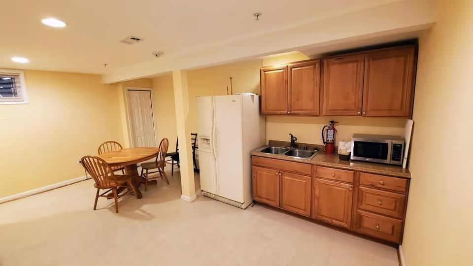 A small kitchenette with wooden cabinets, a refrigerator, sink and microwave beside a round wooden dining table and chairs in a finished room.