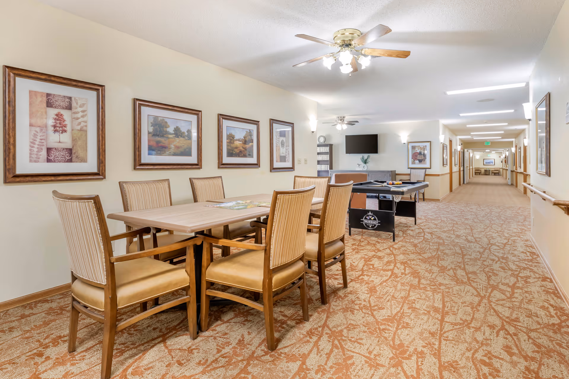 Communal senior living common area with a dining table and chairs, framed artwork on the wall, a game table and TV with a long hallway beyond.