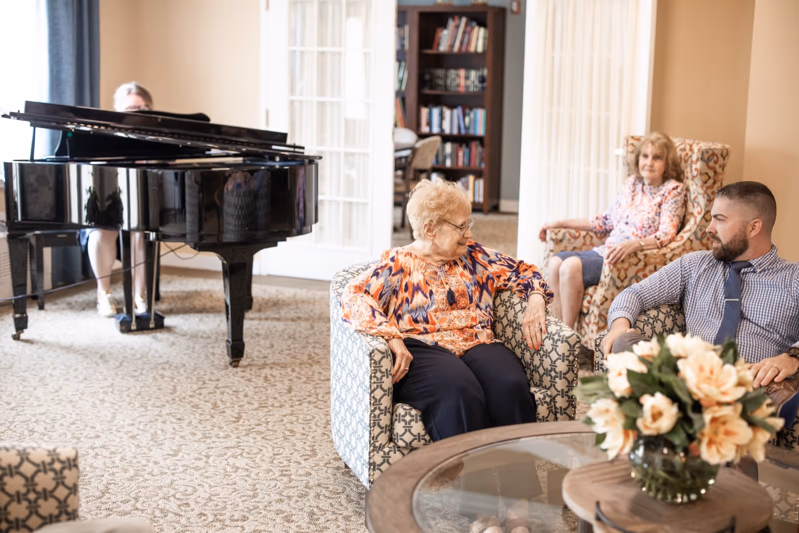 A senior living facility common area with three elderly women and a man sitting on patterned armchairs around a glass coffee table with a flower arrangement. In the background, a person is playing a grand piano near large windows and a bookshelf.