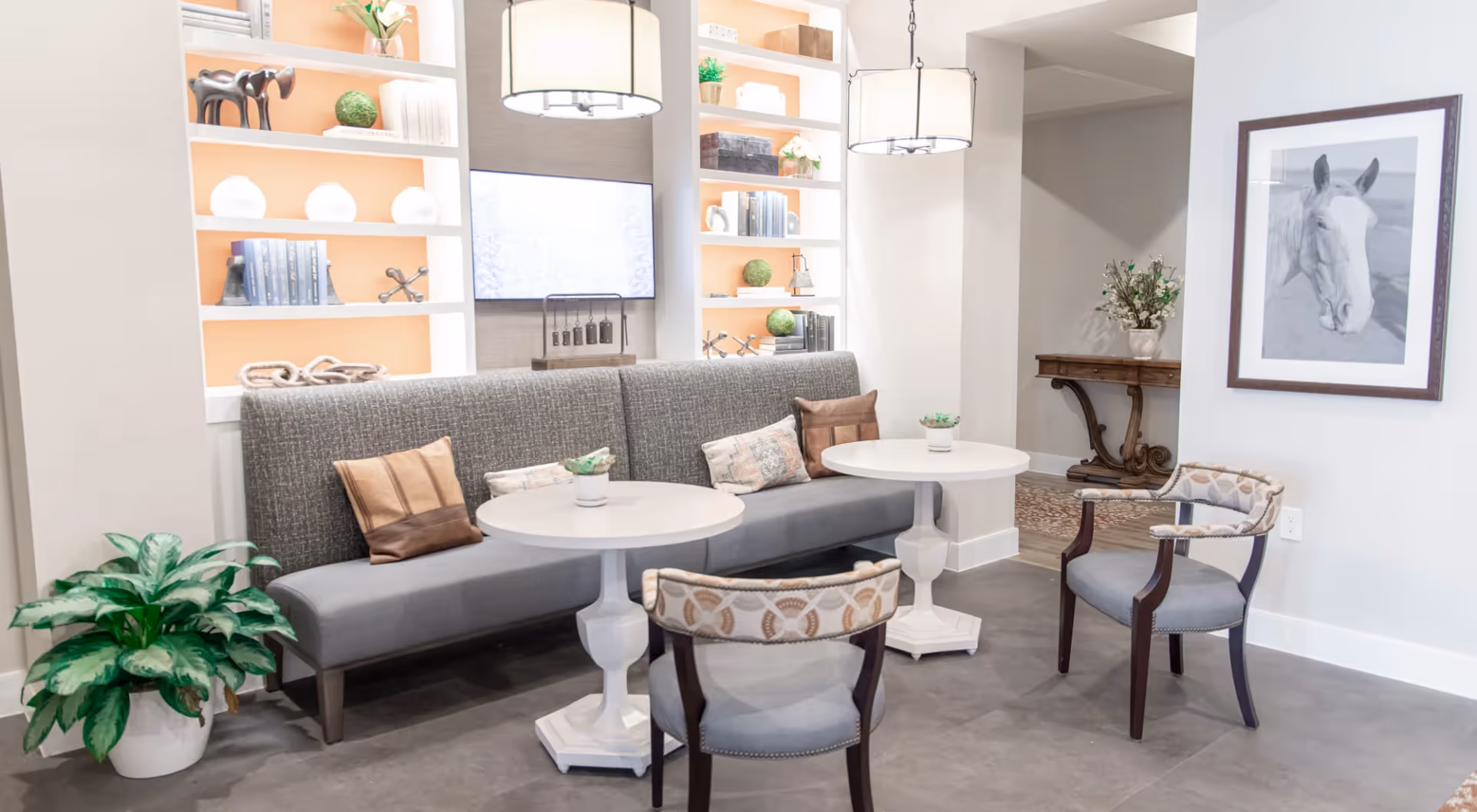 A cozy seating area with a long gray upholstered bench against a wall with built-in shelves displaying decorative items and books. Two round white tables are placed in front of the bench, each accompanied by a patterned armchair. The room features modern pendant lights, a framed black and white horse portrait on the wall, a potted plant on the floor, and a small console table with a floral arrangement in the background.