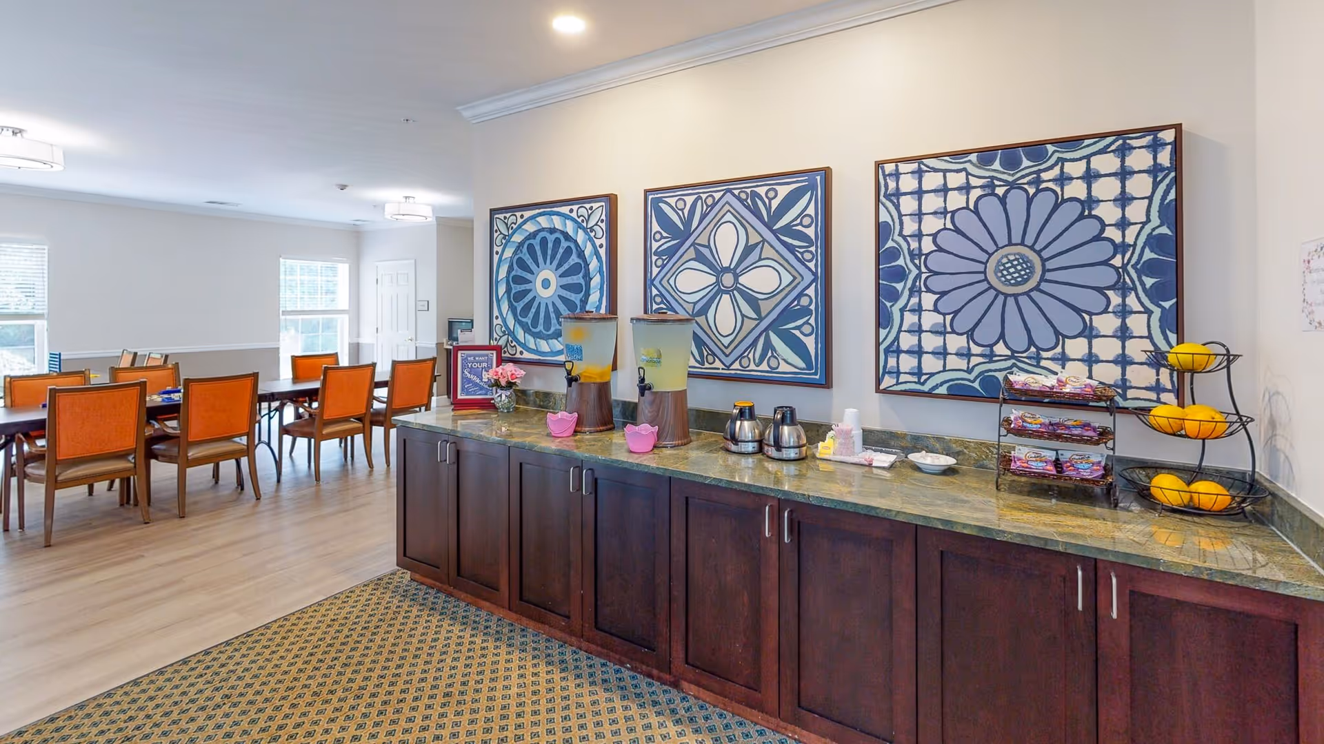 A dining area in a senior living facility with wooden tables and orange cushioned chairs. In the foreground, there is a long dark wooden counter with a green marble top holding two beverage dispensers, coffee pots, snacks, and a three-tier fruit basket with oranges. The wall above the counter features three large framed blue and white floral and geometric patterned artworks. The room has light-colored walls and wood flooring with a patterned carpet near the counter.