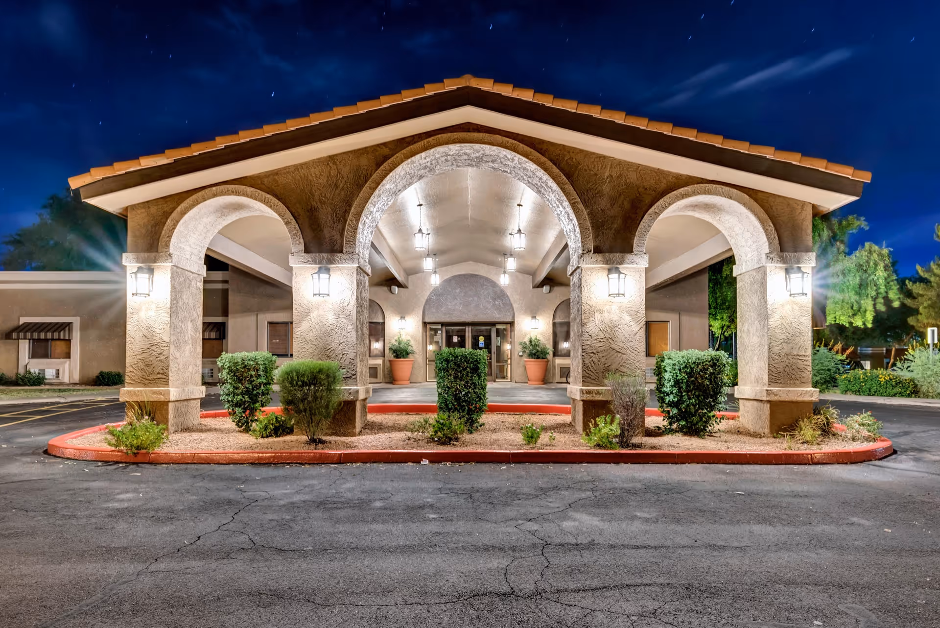 Night view of the front entrance of a building with three large archways supported by columns, illuminated by wall-mounted lanterns and hanging lights. There are small bushes and plants in a landscaped area in front of the entrance, and the sky is dark blue with some clouds.