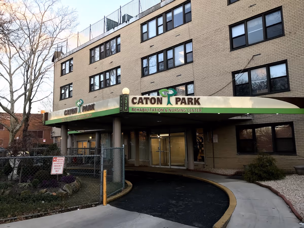 Exterior view of Caton Park Rehabilitation & Nursing Center, a multi-story brick building with a covered entrance and a curved driveway. There are leafless trees and a fenced garden area near the entrance.