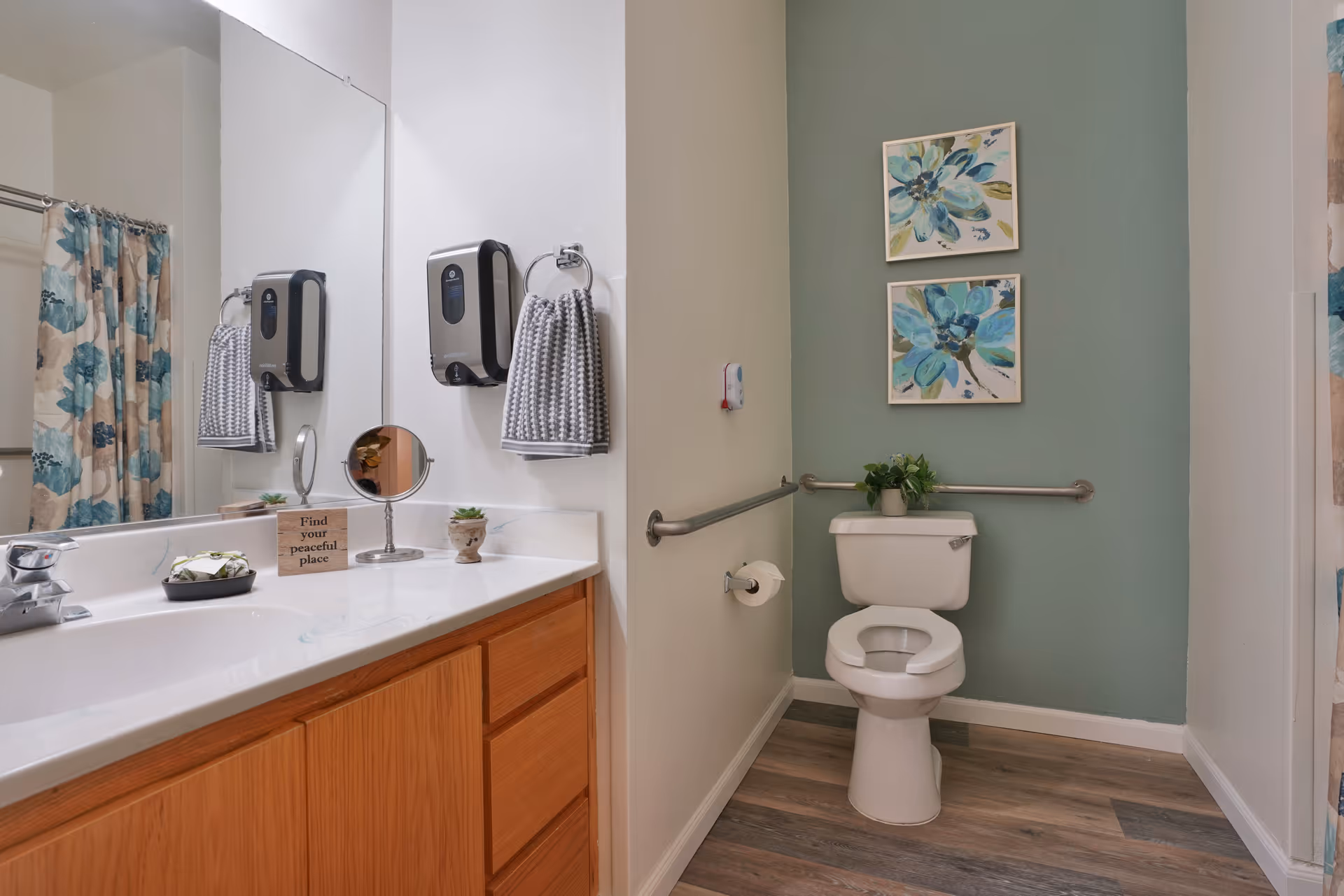 A clean bathroom with a white toilet against a green accent wall decorated with two blue floral paintings. There are metal grab bars on the walls around the toilet. To the left is a wooden vanity with a white countertop, a sink, a small round mirror, a soap dispenser, a gray hand towel, and small decorative plants. A shower curtain with a blue and beige floral pattern is partially visible in the mirror reflection.