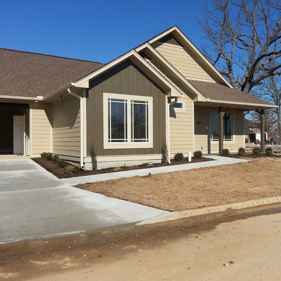 Single-story beige and brown house with a front porch, driveway, and lawn under a clear blue sky.