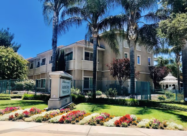 Exterior view of Villas at San Bernardino senior living facility with a well-maintained garden featuring colorful flowers, palm trees, and a white gazebo. The building is a multi-story structure with balconies and surrounded by a fence.