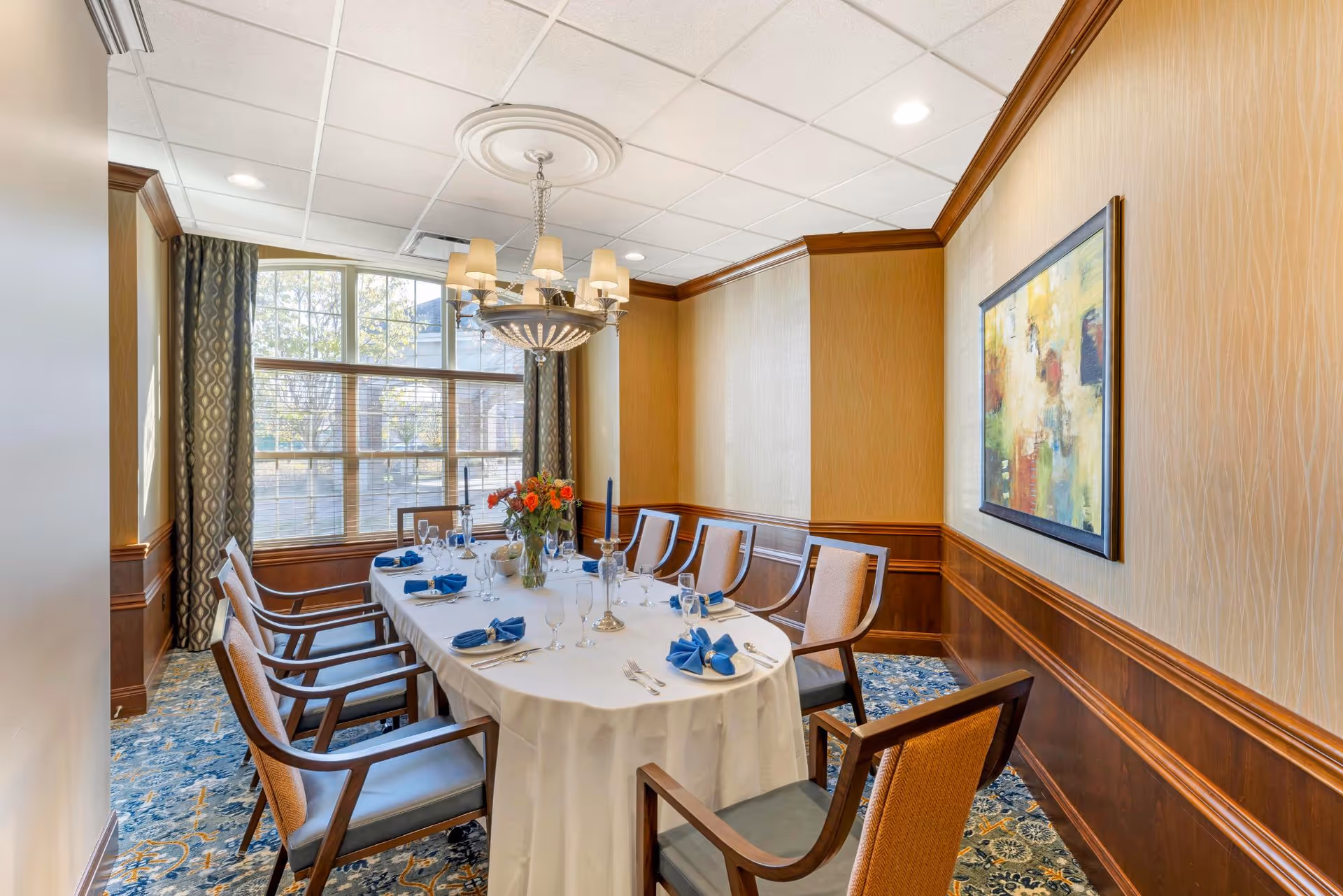 A formal dining room with a long rectangular table covered with a white tablecloth. The table is set with blue napkins, glassware, and silverware. There are eight wooden chairs with cushioned seats around the table. A chandelier hangs above the table, and a large window with patterned curtains lets in natural light. The walls are decorated with wood paneling and a colorful abstract painting.