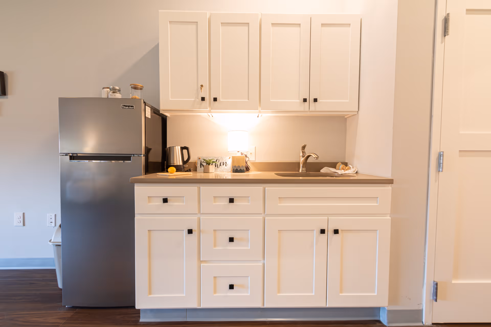 A small kitchen area with white cabinets and drawers, a stainless steel refrigerator, a countertop with a sink, a kettle, a small potted plant, a lemon on a cutting board, and a lamp providing warm light under the upper cabinets.