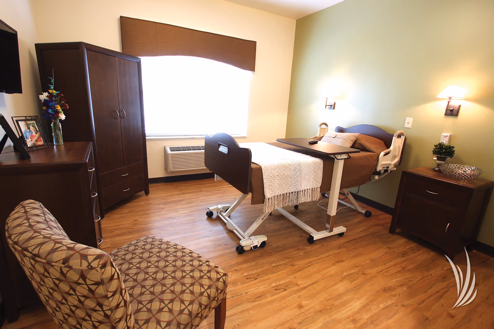 A furnished senior care bedroom with an adjustable hospital bed, bedside tables, dresser, patterned chair and a large window.