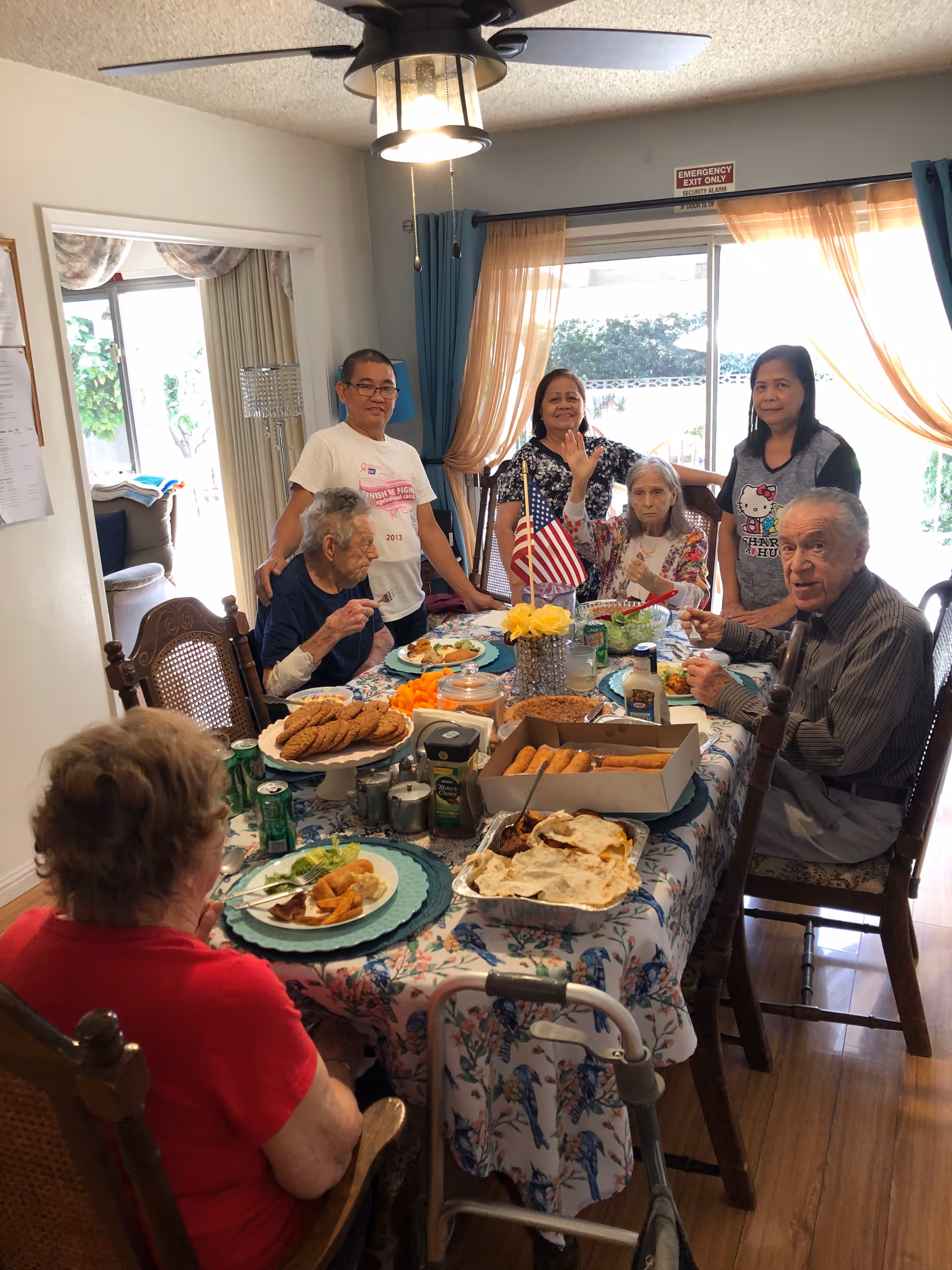 A group of elderly people and two caregivers gathered around a dining table with various foods including cookies, spring rolls, and salad. The room is well-lit with natural light coming through a large window with sheer curtains. An American flag is placed in the center of the table. A ceiling fan with a light fixture is visible above the table.