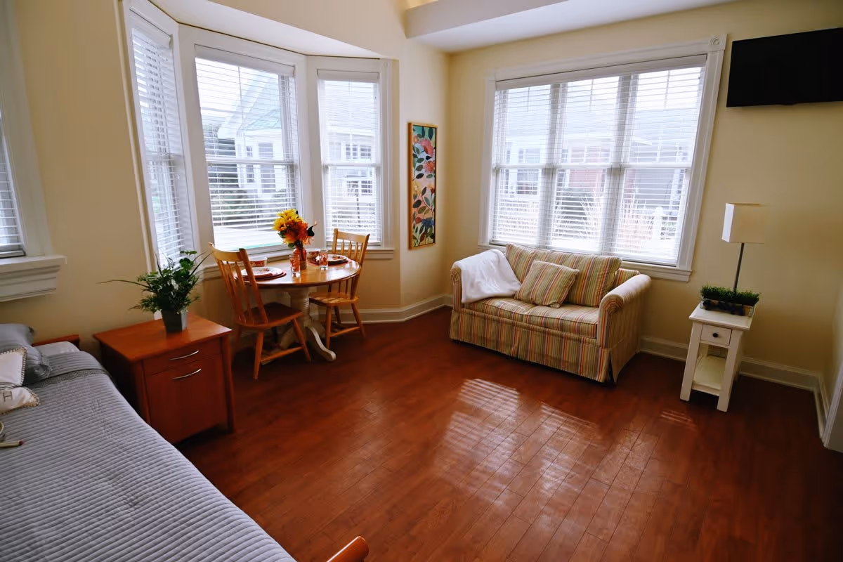 A bright and cozy living area with large windows letting in natural light. The room features a small round wooden dining table with two chairs, a striped loveseat with cushions and a white throw, a wooden side table with a potted plant, a floor lamp, and a mounted flat-screen TV. The floor is hardwood, and the walls are painted a soft cream color.