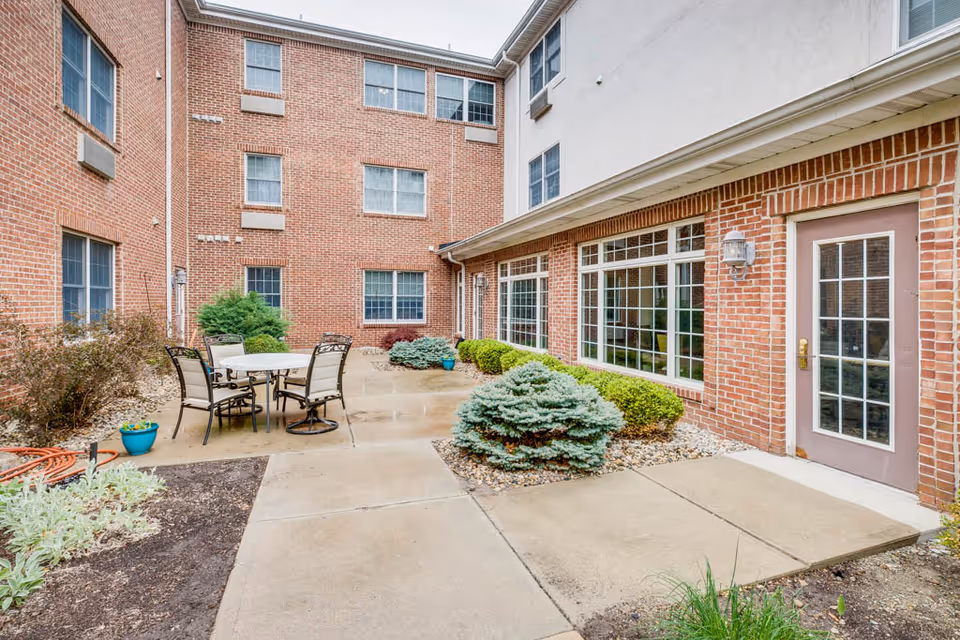 Outdoor courtyard area of a brick building with a concrete pathway, patio table with four chairs, various shrubs and plants, and large windows along the building wall.
