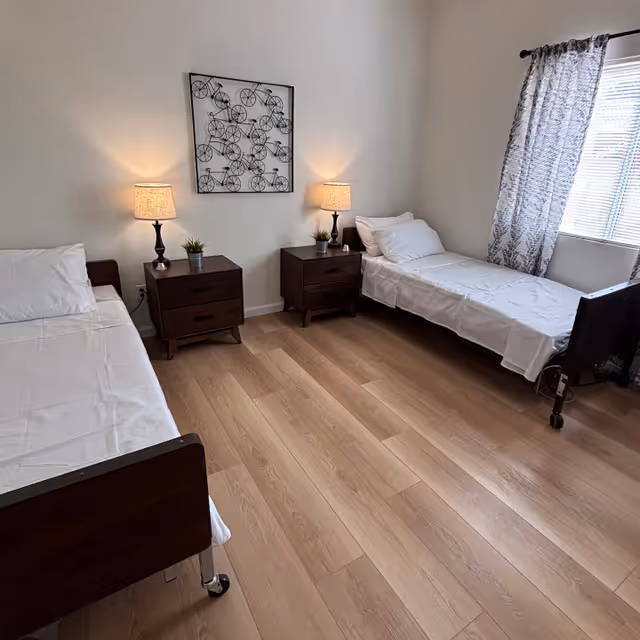 A simple bedroom with two single beds on opposite sides of the room, each with white bedding. Between the beds are two dark wooden nightstands, each with a lamp and a small potted plant. A window with patterned curtains lets in natural light on the right side. A decorative wall art featuring multiple bicycles is hung above the nightstands. The floor is light wood.