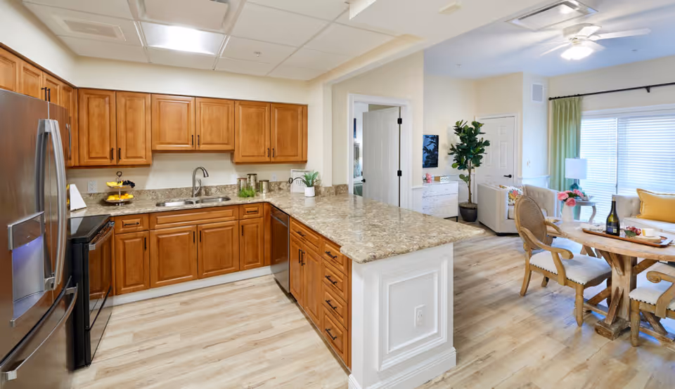 A bright and spacious kitchen with wooden cabinets, granite countertops, a stainless steel refrigerator, oven, and dishwasher. The kitchen opens into a dining area with a wooden table and chairs, a living room with a sofa, a large window with green curtains, and a ceiling fan. The flooring is light wood, and there are decorative plants and flowers adding a cozy touch.