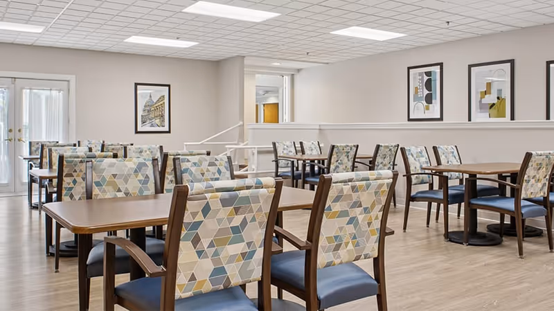 A bright dining room in a senior living community with multiple wooden tables and chairs featuring geometric patterned upholstery. The room has light-colored walls, framed artwork, and large windows allowing natural light to enter.