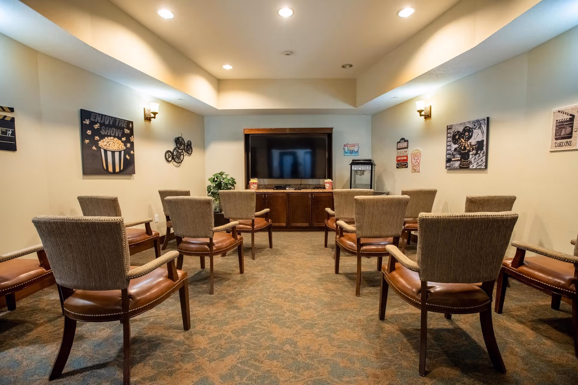 A small theater-style room with two rows of chairs facing a flat-screen TV mounted on a wooden cabinet. The walls are decorated with movie-themed artwork and there is a popcorn machine on the right side of the TV cabinet. The room has soft lighting from wall sconces and a carpeted floor.