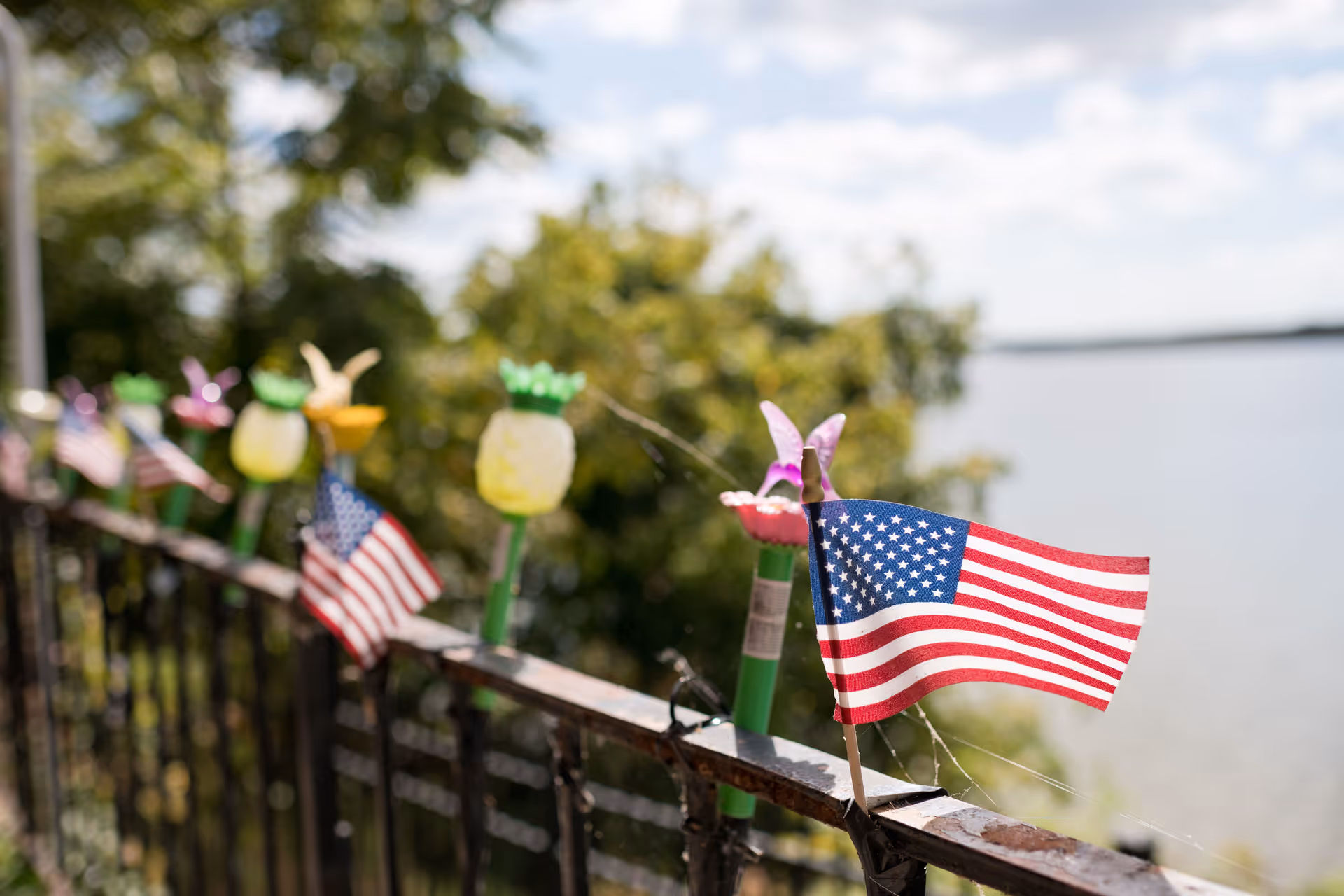 Close-up view of a metal railing decorated with small American flags and colorful flower-shaped solar lights, with a blurred background of trees and a body of water under a partly cloudy sky.