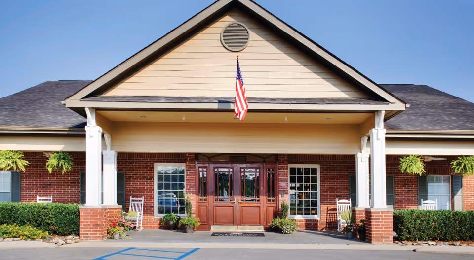 Front exterior view of a single-story brick building with a covered entrance supported by white columns. An American flag hangs above the entrance, which features double wooden doors with glass panels. There are windows on either side of the doors, white rocking chairs, hanging plants, and neatly trimmed bushes along the front.