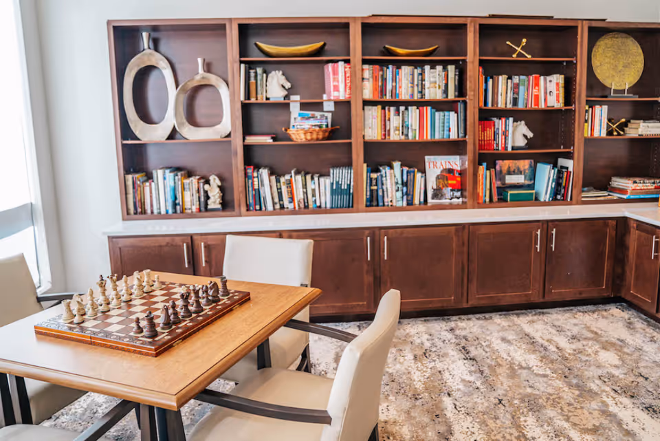 A cozy room with a wooden table set for a chess game, surrounded by four cushioned chairs. Behind the table is a large wooden bookshelf filled with books and decorative items, including sculptures and bowls. The room has a patterned carpet and natural light coming from a window on the left.