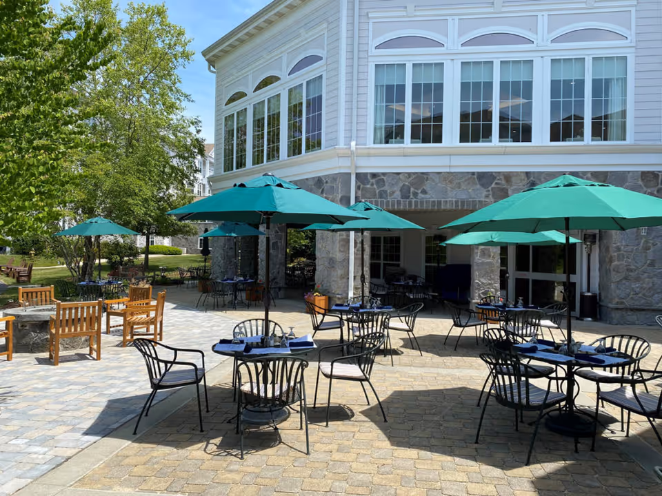 Outdoor patio area at StoneRidge Senior Living with several tables and chairs under green umbrellas, surrounded by trees and a stone building with large windows in the background.