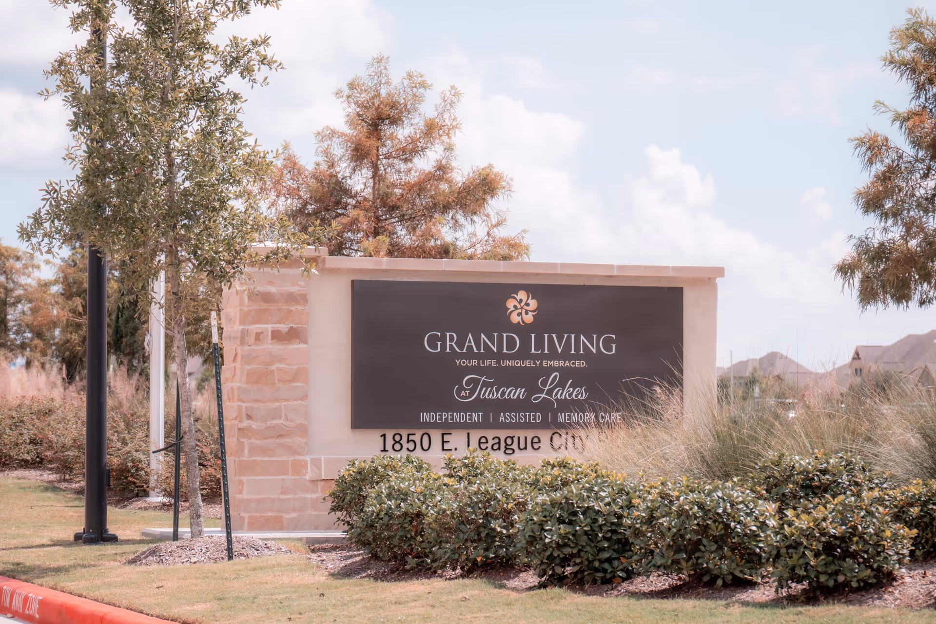 Outdoor view of a stone and stucco sign for Grand Living at Tuscan Lakes, surrounded by bushes and trees under a partly cloudy sky.