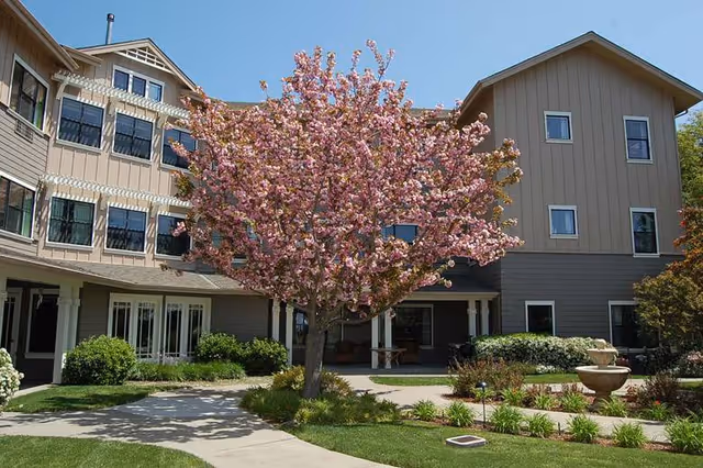 Exterior view of a senior living facility with a large pink flowering tree in the center, surrounded by well-maintained landscaping, walkways, and a multi-story building with beige and gray siding and multiple windows.