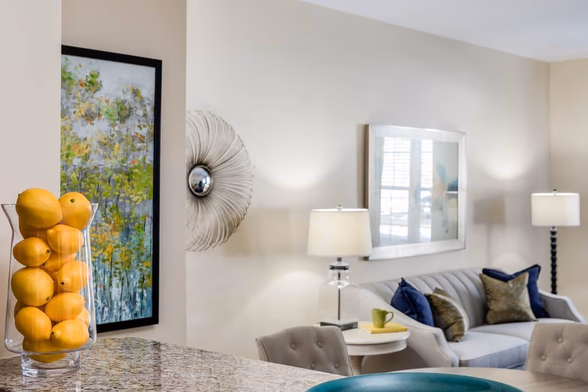 A modern living room with a gray sofa adorned with blue and brown pillows, two table lamps on either side, a round side table with a green mug and a book, a decorative wall mirror, and a framed abstract painting. In the foreground, a glass vase filled with lemons sits on a granite countertop.