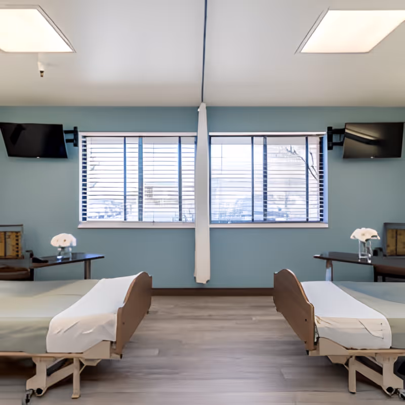 A room with two hospital-style beds positioned parallel to each other, each with a white sheet and light green bedding. Between the beds are small tables with white flowers in glass vases. The room has light blue walls, large windows with blinds, and two wall-mounted flat-screen televisions above each bed.