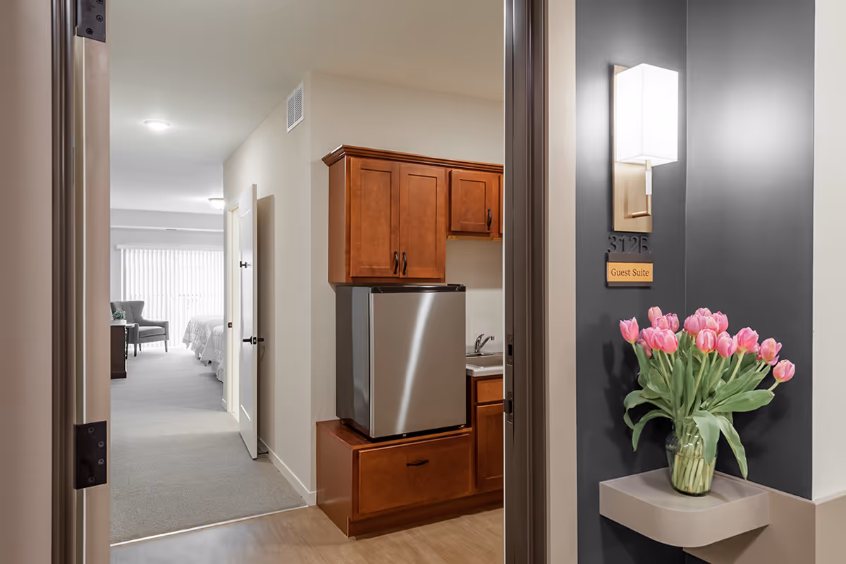 View into a guest suite labeled 312B at Lexington Landing, showing a small kitchenette with wooden cabinets and a mini refrigerator, a hallway leading to a bedroom area with a bed and chair near a window with vertical blinds, and a wall-mounted light fixture above a shelf holding a vase of pink tulips.