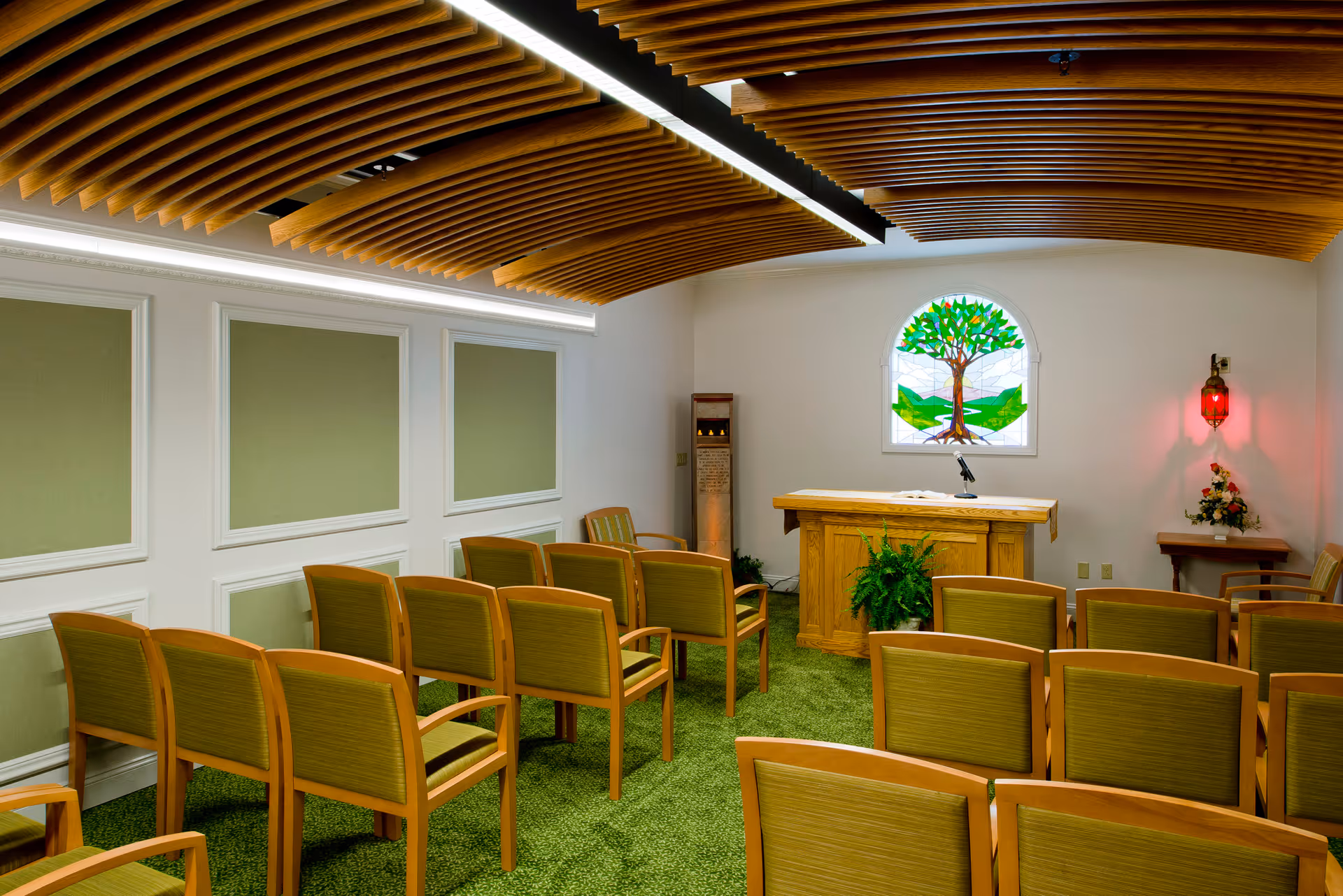 Interior view of a small chapel or meditation room with rows of wooden chairs with green cushions facing a wooden podium. The room has a green carpet, a stained glass window depicting a tree, and a wooden slatted ceiling with modern lighting. There is a small table with flowers and a red lamp on the right side.