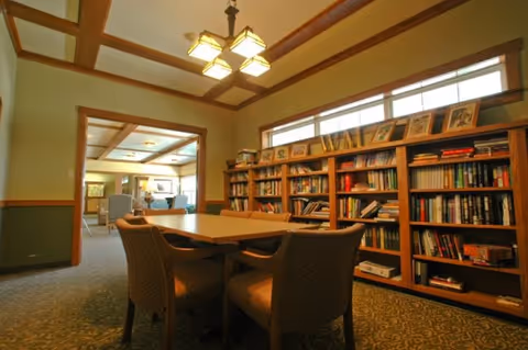 A cozy interior room with a wooden table surrounded by four upholstered chairs. There is a large bookshelf filled with books and framed pictures along one wall. The room has a carpeted floor, wooden trim, and a ceiling light fixture with multiple lamps. An open doorway leads to another room with additional seating and natural light.