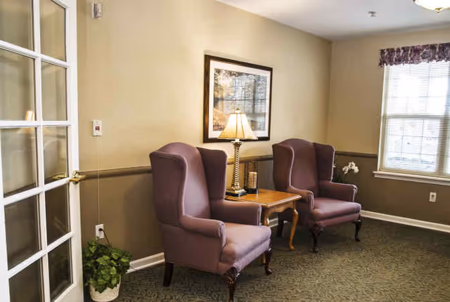 A cozy sitting area in a senior living facility featuring two purple wingback chairs separated by a wooden side table with a lamp and decorative items. The room has beige walls, a framed picture, a window with floral valance, and a potted plant near a glass-paneled door.