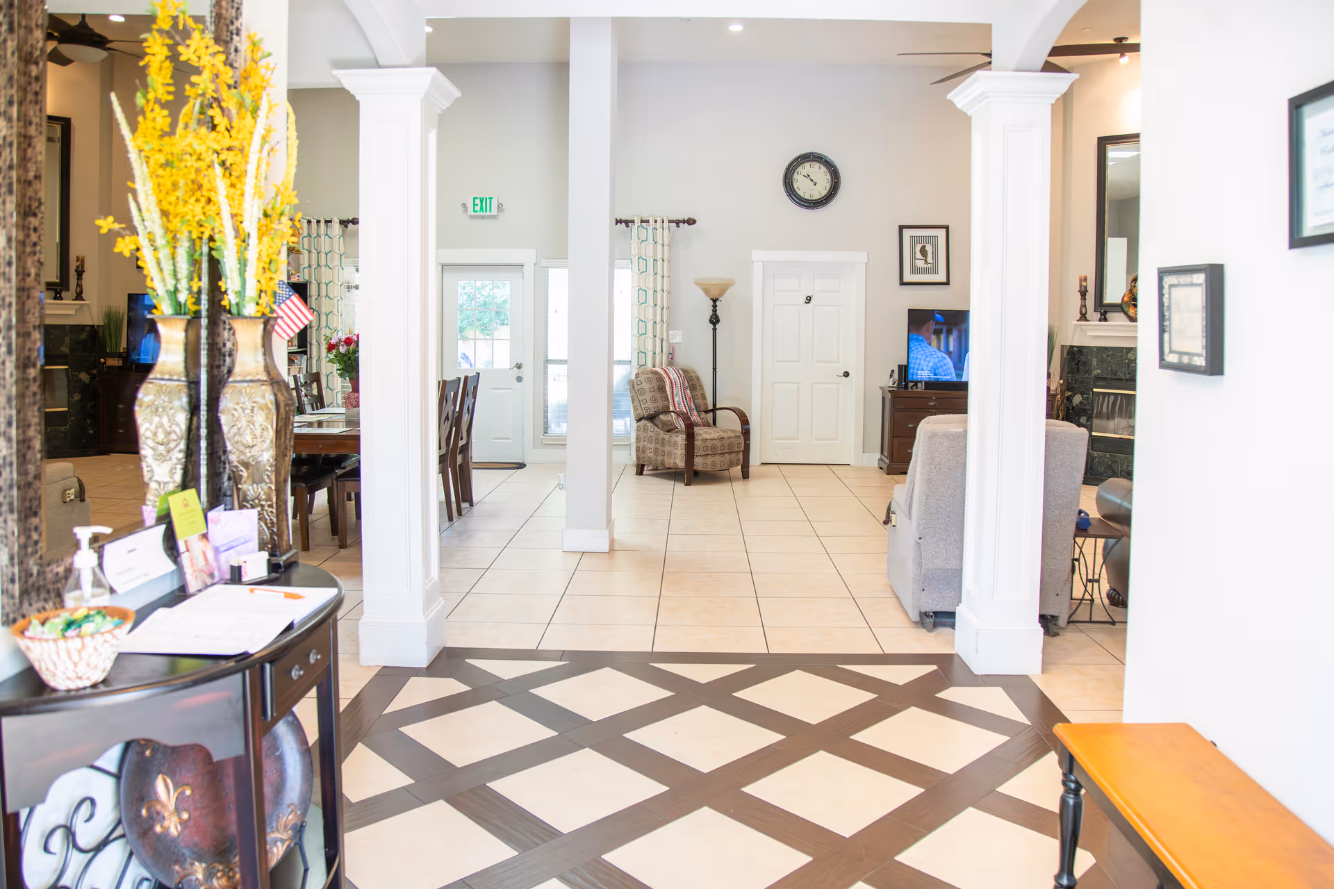 Bright and spacious interior of a senior living facility featuring a tiled floor with a dark wood pattern, white columns, a small table with a large vase of yellow flowers, a dining area with a table and chairs, a sitting area with armchairs, a wall clock, and framed pictures on the walls.