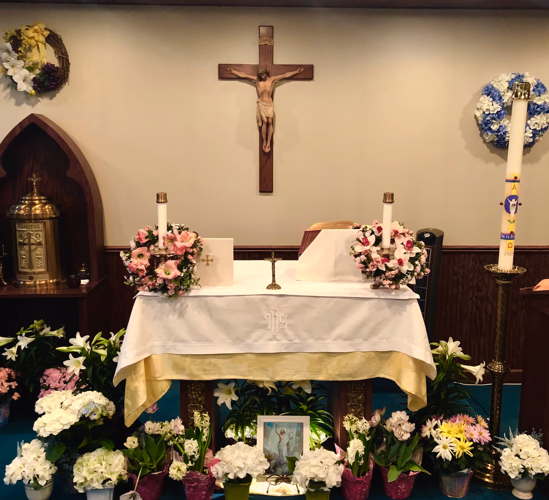 A religious altar decorated with a white and gold cloth, two floral candle holders with pink and white flowers, a small crucifix in the center, and a framed picture of Jesus surrounded by various potted flowers. A wooden crucifix with Jesus is mounted on the wall behind the altar. To the right, there is a tall decorated candle on a brass stand, and to the left, a brass container with religious symbols inside a wooden alcove. Floral wreaths are hung on the wall on both sides of the crucifix.