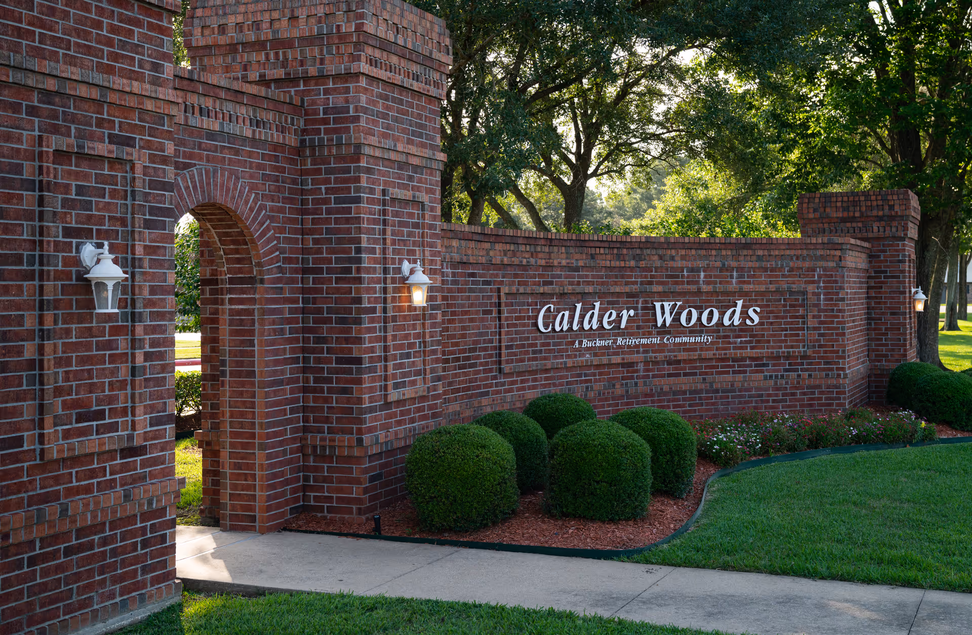Brick entrance sign for Calder Woods, a Buckner Retirement Community, surrounded by neatly trimmed bushes and trees in the background.