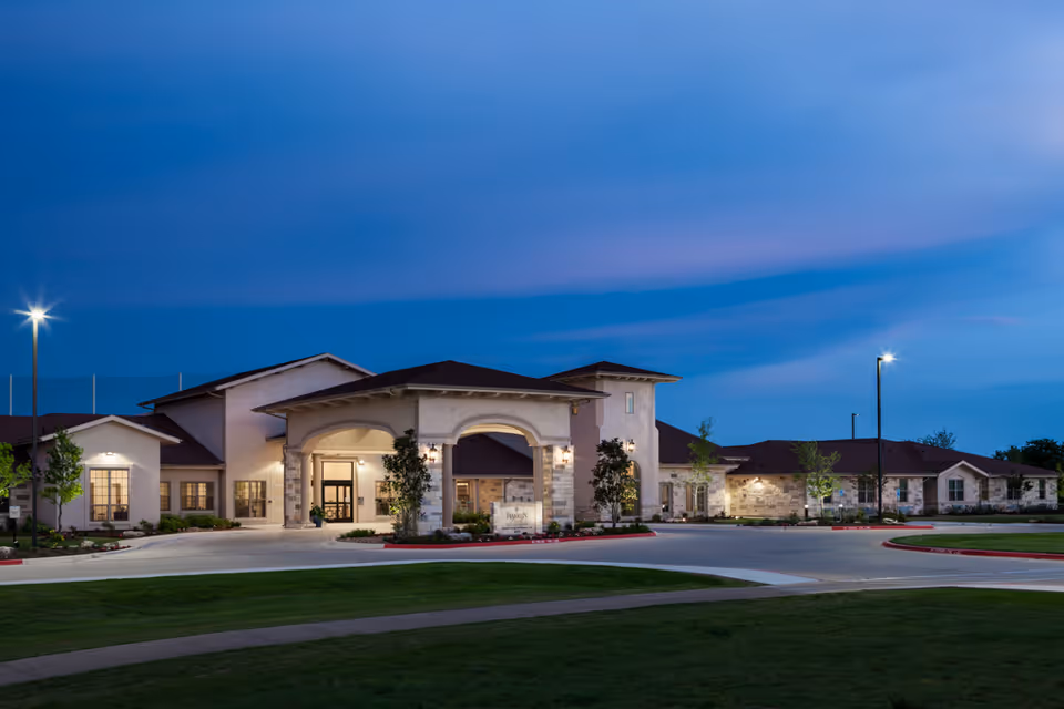 Exterior view of Volante Senior Living of Round Rock building at dusk with illuminated entrance, surrounding greenery, and street lamps.