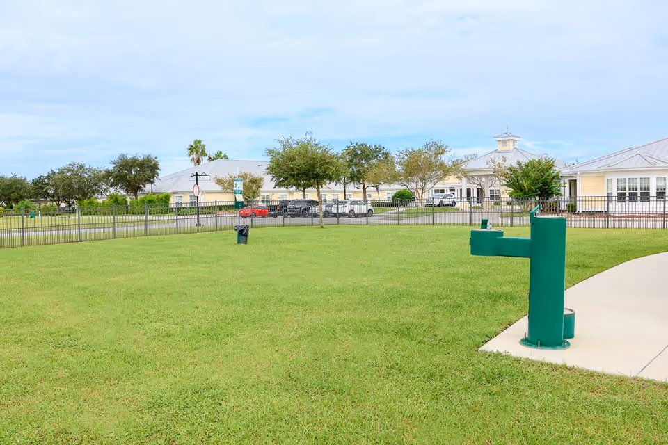 A grassy outdoor area with a green water fountain and a paved walkway. In the background, there is a fenced senior living facility building with several trees and parked cars under a partly cloudy sky.
