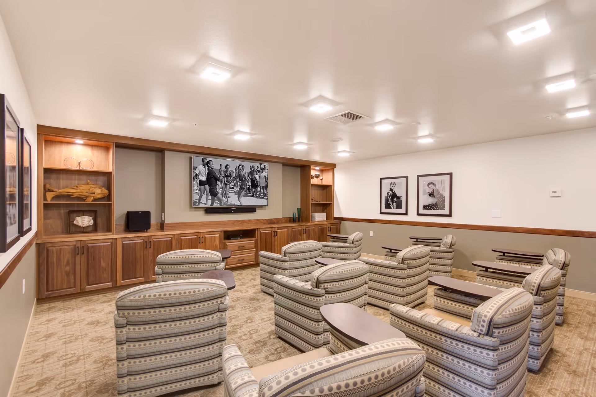A cozy media room with multiple striped armchairs arranged in rows facing a large flat-screen TV mounted on a wooden built-in cabinet. The room has soft lighting from ceiling fixtures, framed black and white photos on the walls, and a carpeted floor.