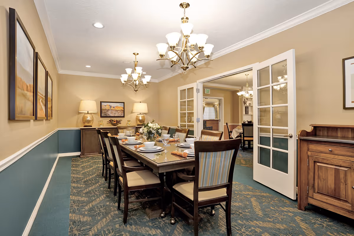 A well-lit dining room with a long wooden table set for a meal, including plates, bowls, glasses, and napkins. The room features beige walls with a blue wainscoting, patterned carpet, two chandeliers, framed artwork on the walls, and a wooden sideboard. French doors lead to another room with similar decor.