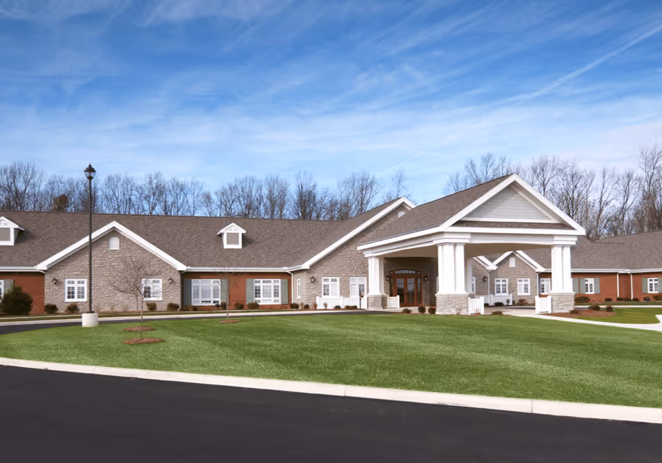 Exterior view of The Inn at Ashland Woods, a single-story building with a covered entrance supported by white columns, surrounded by a well-maintained lawn and a paved driveway. Leafless trees are visible in the background under a partly cloudy blue sky.