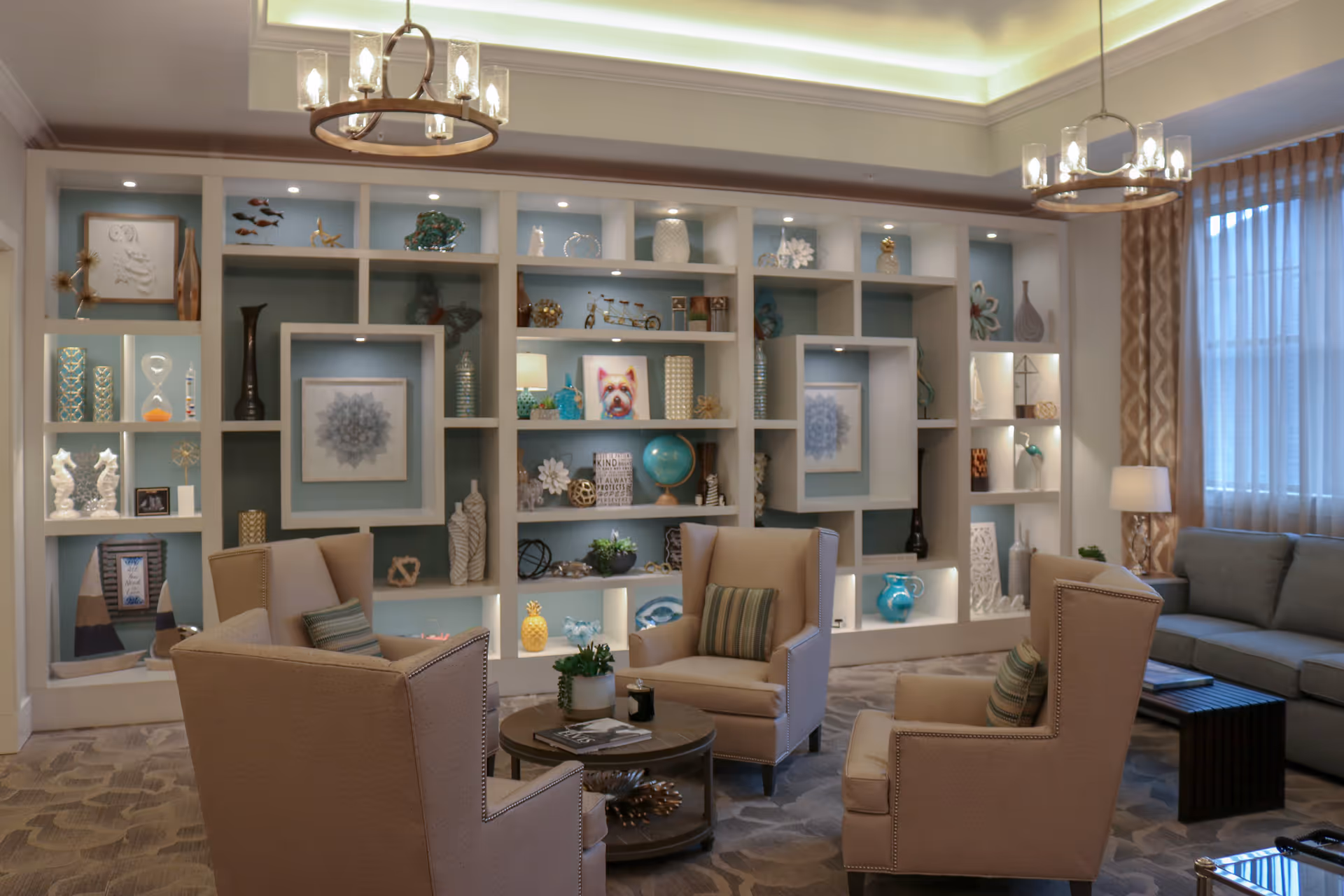 A cozy living room area with four beige armchairs arranged around a round wooden coffee table. Behind the chairs is a large built-in shelving unit filled with decorative items such as vases, sculptures, framed artwork, and a globe. The room has soft lighting from two chandeliers and a table lamp near a window with sheer curtains. A gray sofa is visible on the right side.