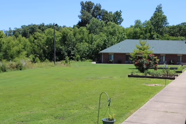 A grassy outdoor area with a concrete walkway on the right side leading to a single-story brick building with a green roof. There are trees and bushes in the background under a clear blue sky.