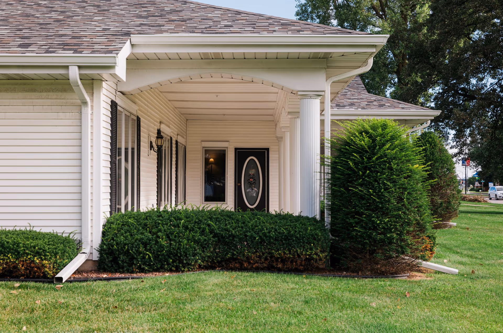 Front porch of a single-story building with white siding, a covered entrance supported by white columns, a black door with an oval glass panel, and neatly trimmed green bushes and lawn in the foreground.