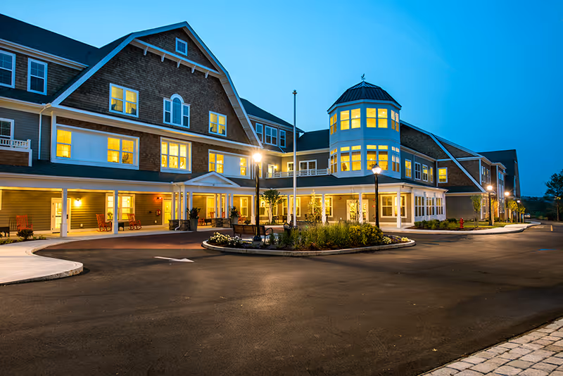 Exterior view of a large multi-story senior living facility building at dusk with lights glowing warmly from the windows. The building features a covered entrance, a roundabout driveway, landscaped greenery, and several lampposts illuminating the area.