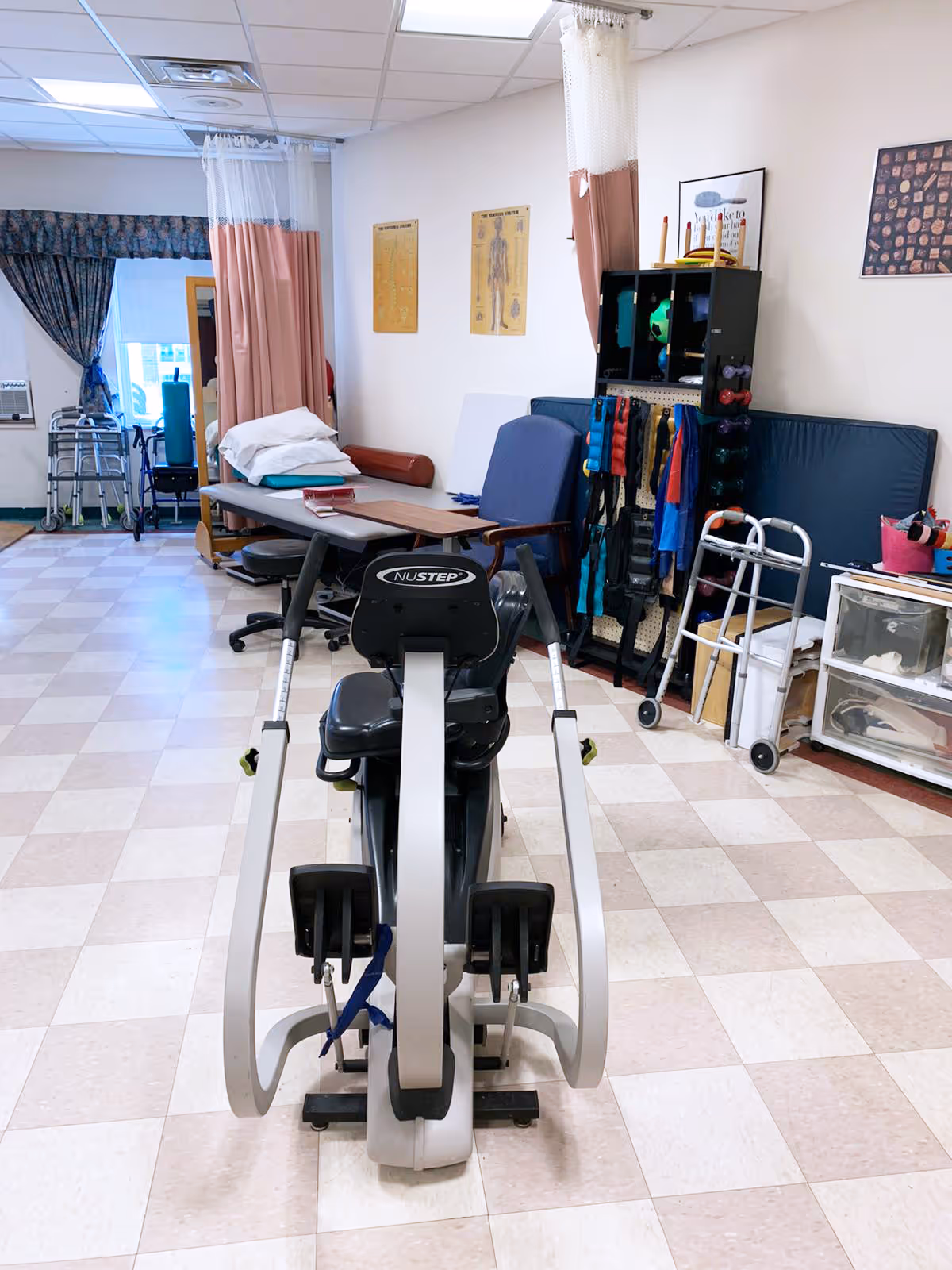 Physical therapy room with exercise equipment including a Nustep machine in the foreground, a treatment table with pillows, chairs, walkers, and various therapy tools and supplies organized on shelves and hanging on the wall.